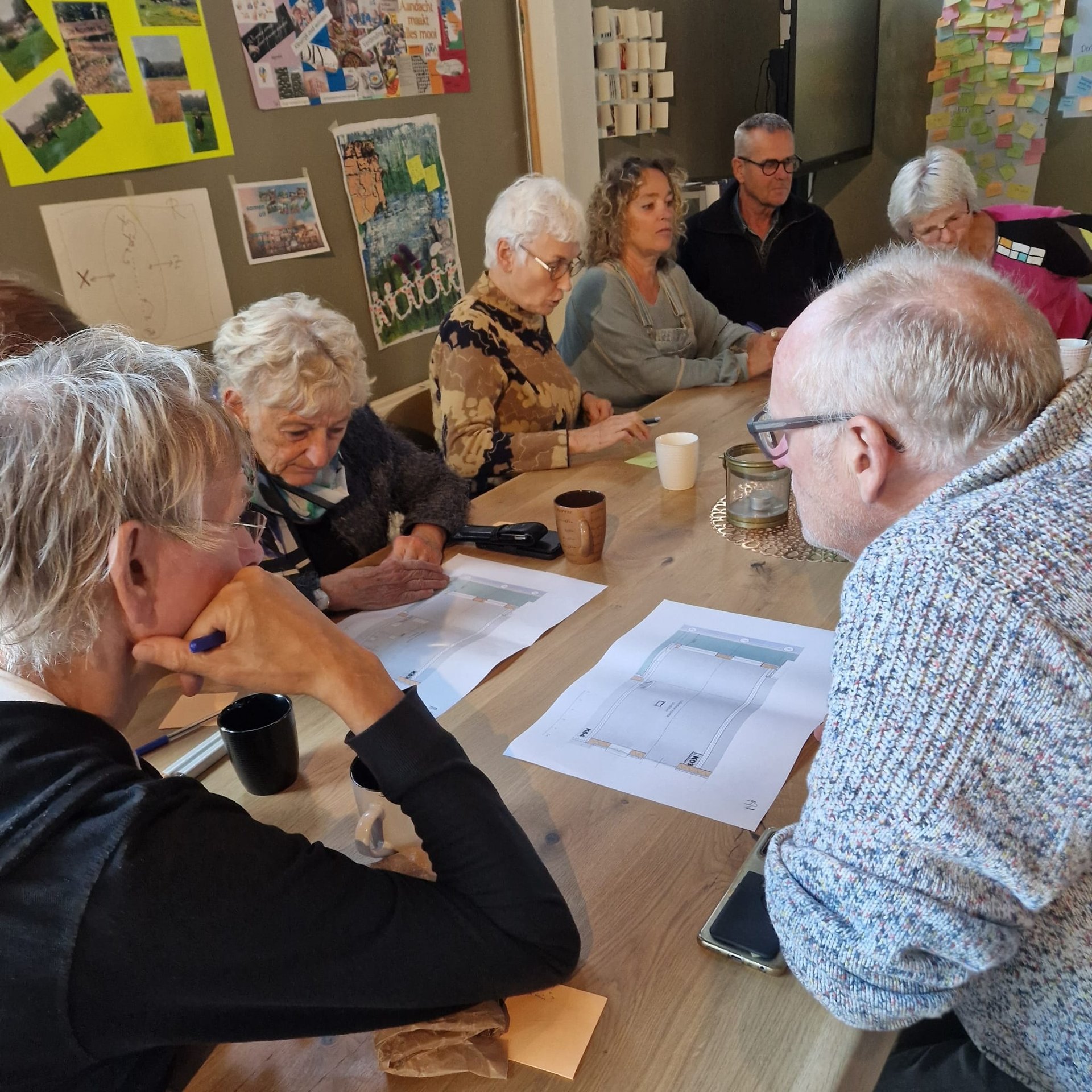Vijf personen zitten rondom een houten tafel met daarop uitgeprinte documenten. Ze zijn met elkaar in gesprek.